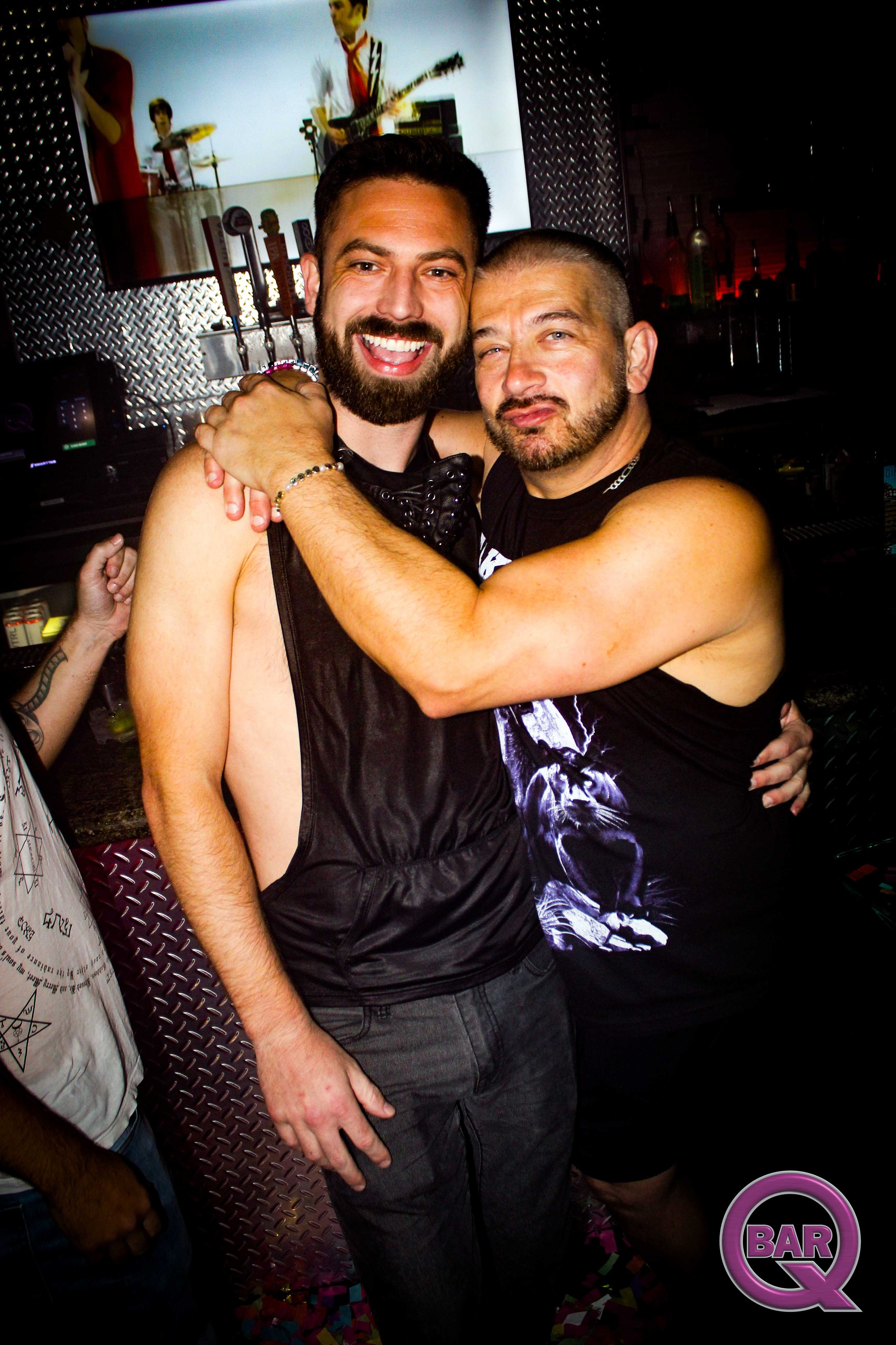 Two men hugging and smiling at a party in a bar or club. One man has a beard and short dark hair, wearing a black tank top. The other man has a buzz cut and a beard, wearing a black graphic t-shirt. There are disco confetti on the floor and a large s