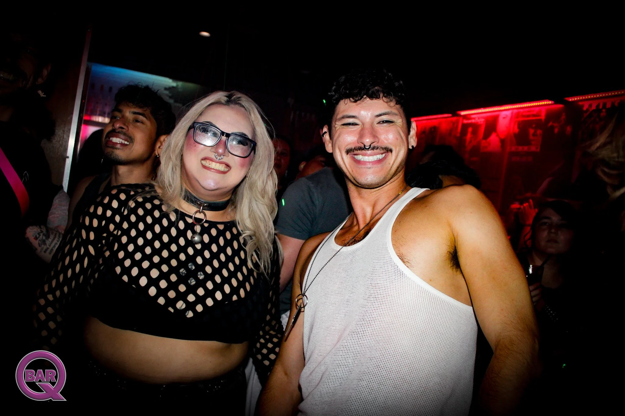 Two smiling people taking a selfie in a bar, with a woman wearing glasses and a man in a white tank top, surrounded by others and red lighting.