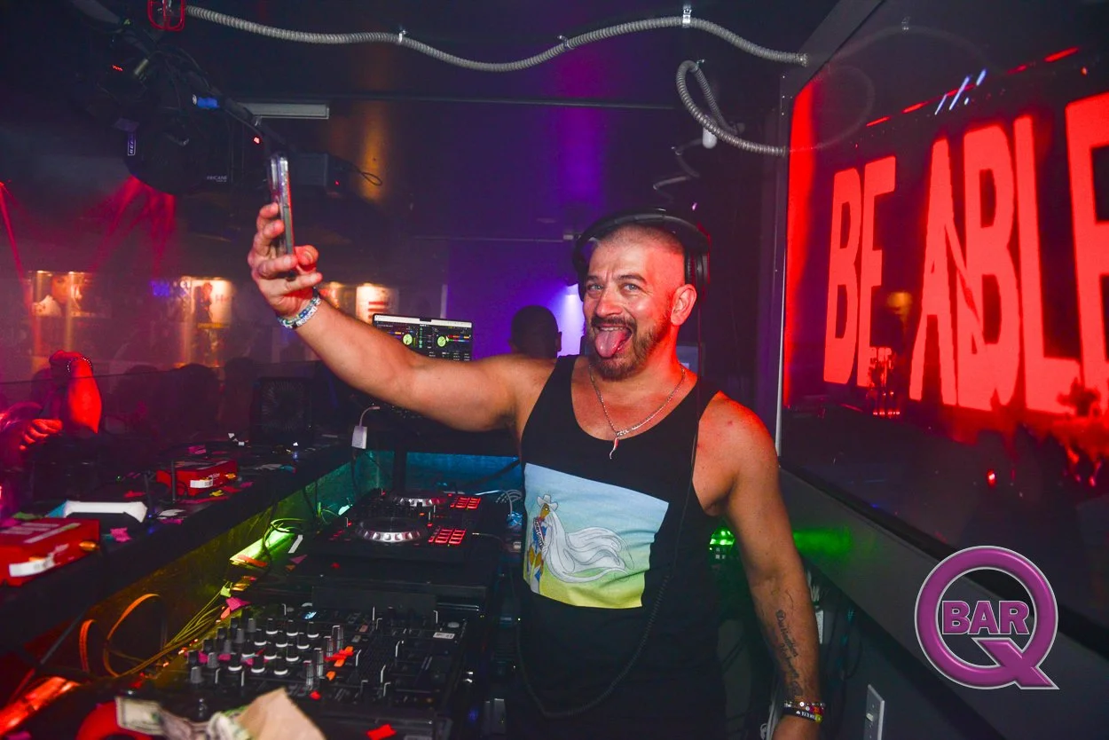 A man with a beard and tattoos, wearing a black tank top with a unicorn on it, is taking a selfie while DJing at a nightclub. He is smiling and sticking his tongue out, with headphones on his head. There is a large red sign that says 'BE ANDLE' on th