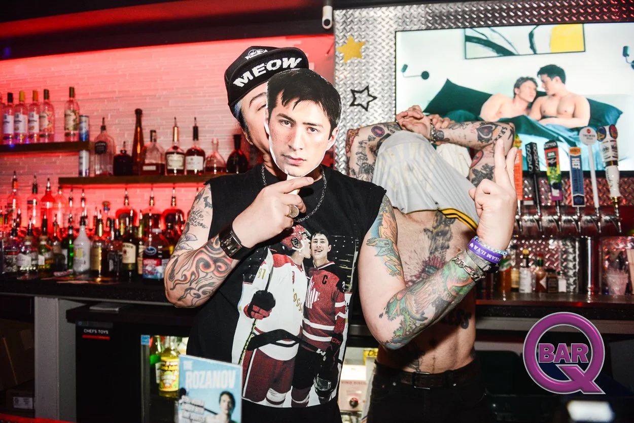 Person with tattoos holding a mask of a man's face in a bar with alcohol bottles and a TV screen in the background showing two shirtless men on a bed.