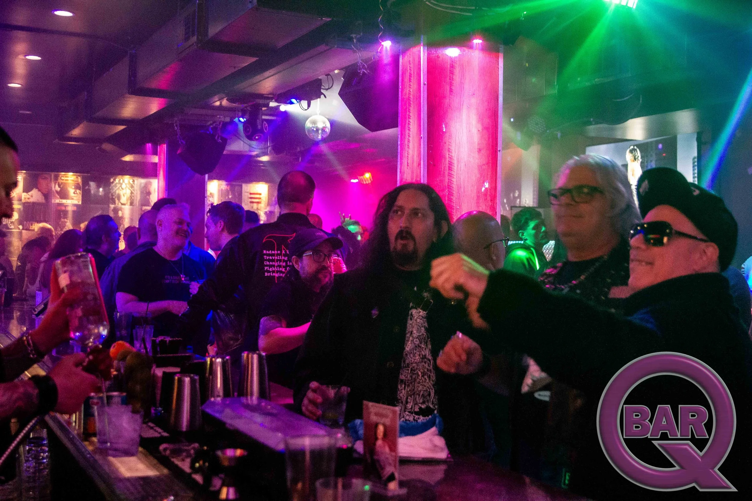 A lively nightclub scene with people dancing and socializing under colorful lighting. A bar area is visible with drinks and a bartender in the foreground. A small disco ball hangs from the ceiling, and there is a neon sign that says 'BAR'.