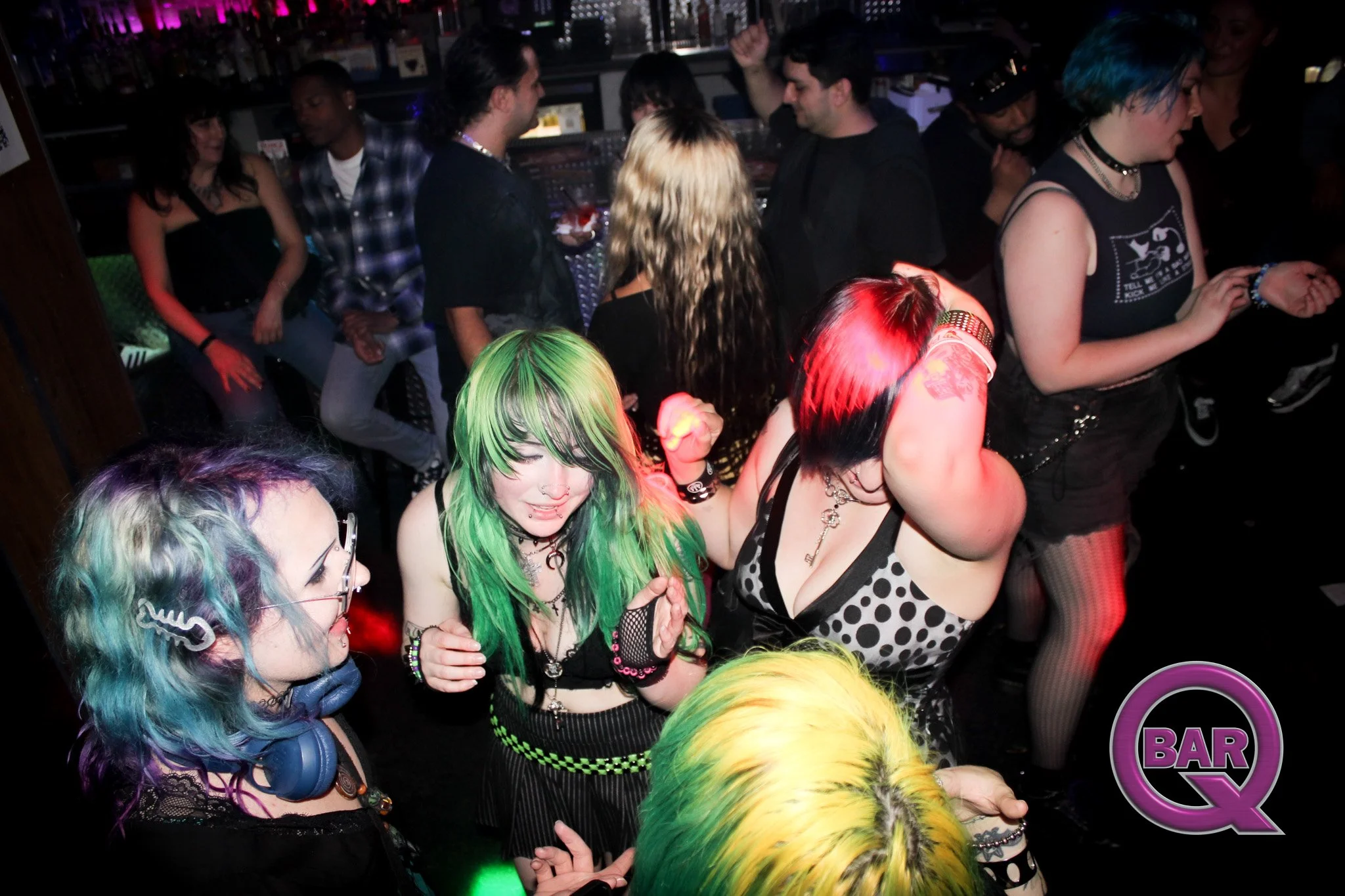Group of young adults dancing and socializing at a nightclub, with colorful hair, tattoos, and punk/goth style clothing, illuminated by neon and stage lighting.
