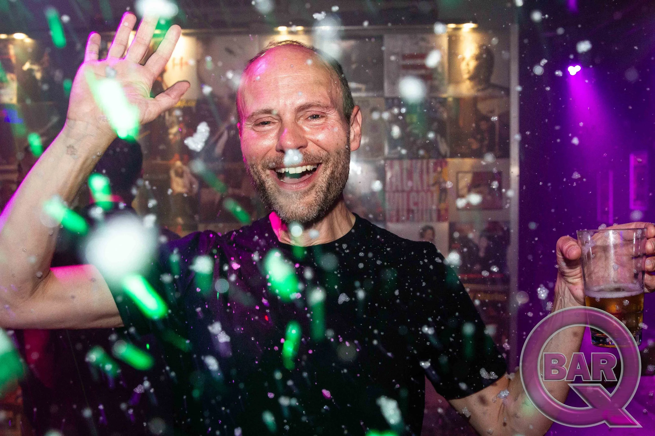 A man smiling and waving behind a glass wall with colorful confetti, holding a glass of beer in a bar.
