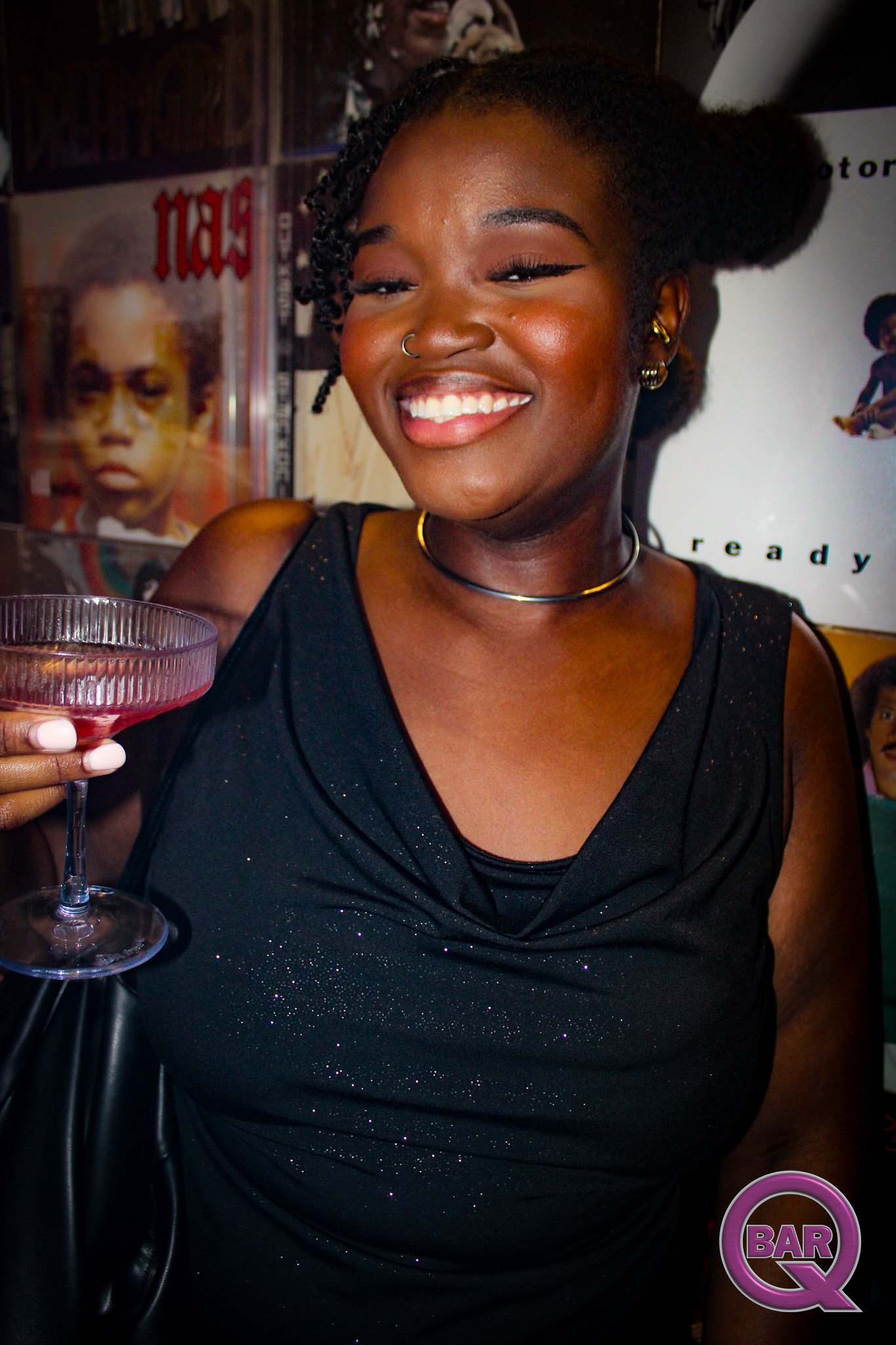 A woman smiling while holding a cocktail glass, wearing a black dress with sparkles, gold jewelry, and a nose ring, at a bar or club setting, with posters and a purple 'Bar Q' logo in the background.