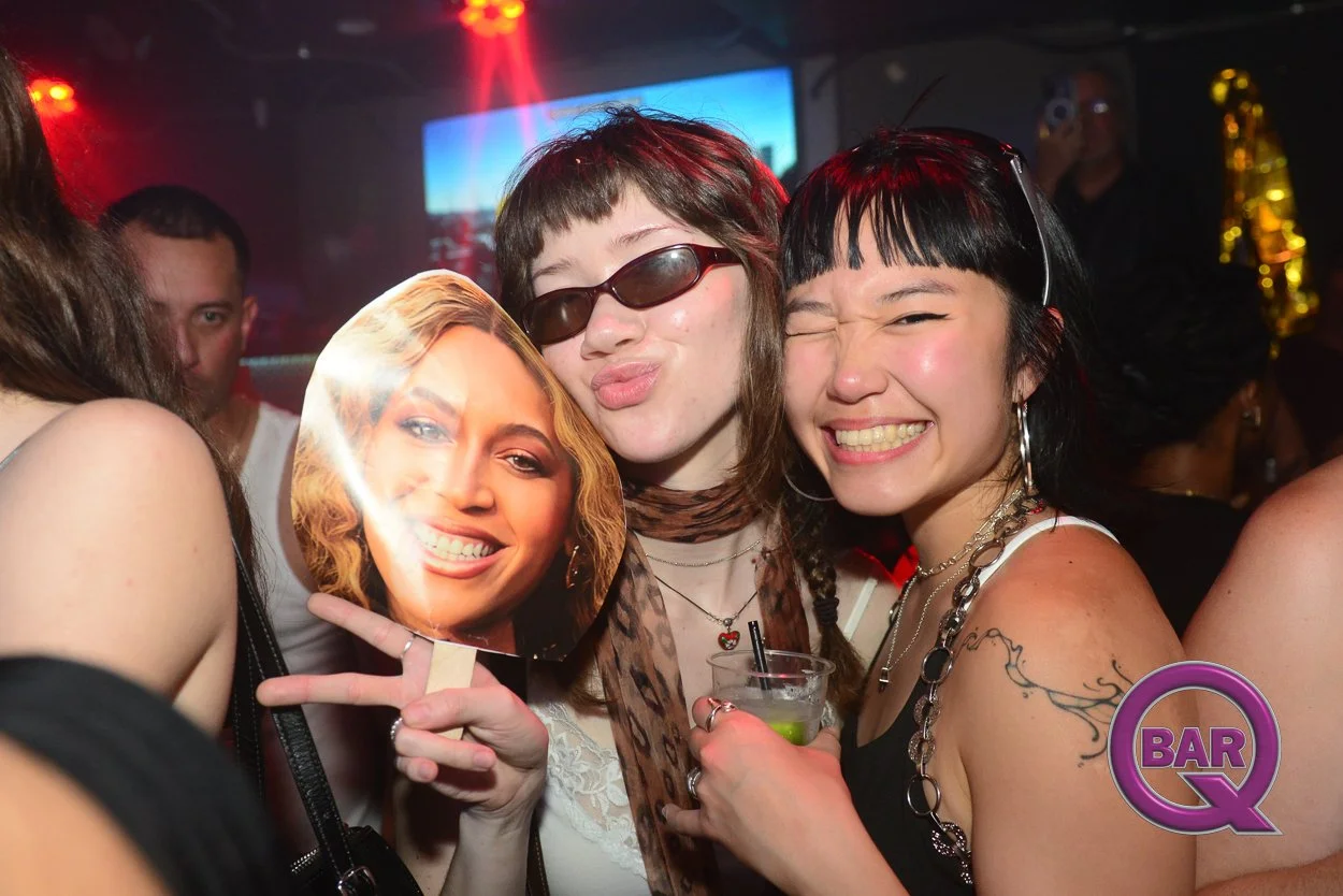 Two women at a bar or nightclub, smiling and posing for the camera, one holding a cutout of a woman's face on a stick, with others in the background enjoying the evening.