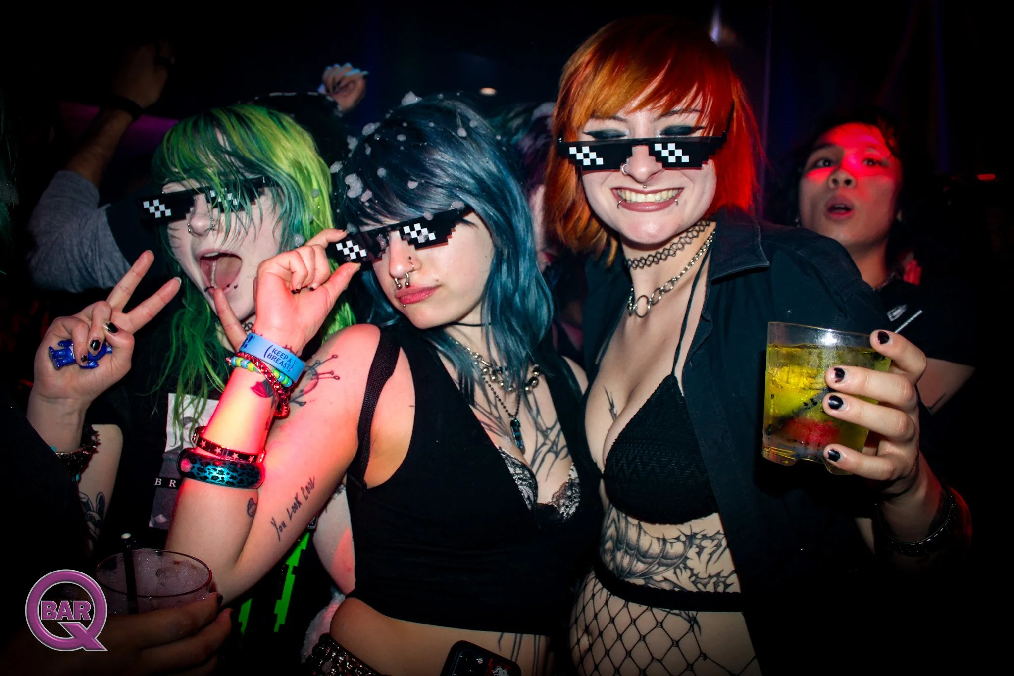 Group of young women at a party wearing pixelated sunglasses, displaying tattoos, piercings, and colorful hair, holding drinks, and enjoying the moment.
