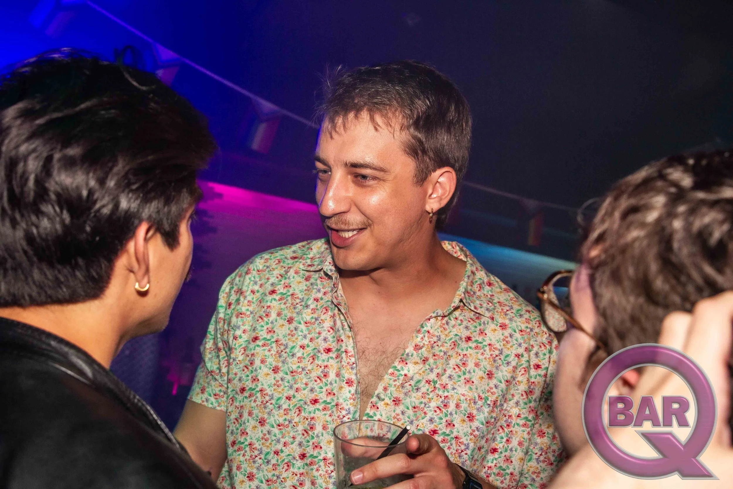 A man with short hair, wearing a floral shirt, is smiling and talking to two women at a bar or nightclub. One woman has dark hair and is wearing earrings. The other woman, partially visible, has dark hair and is holding a drink. The background is dar