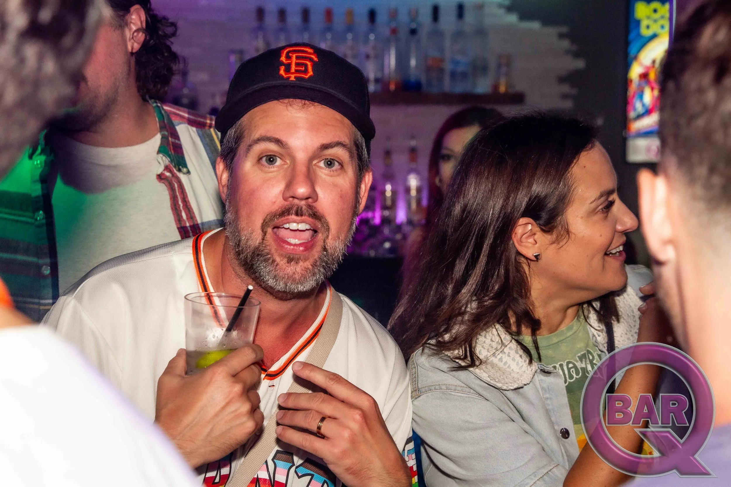 A group of people, including a man with a beard wearing a San Francisco Giants cap, is gathered at a bar. The man is holding a drink with a straw, and there are women and other men in the background, some holding drinks. The bar area has bottles and 