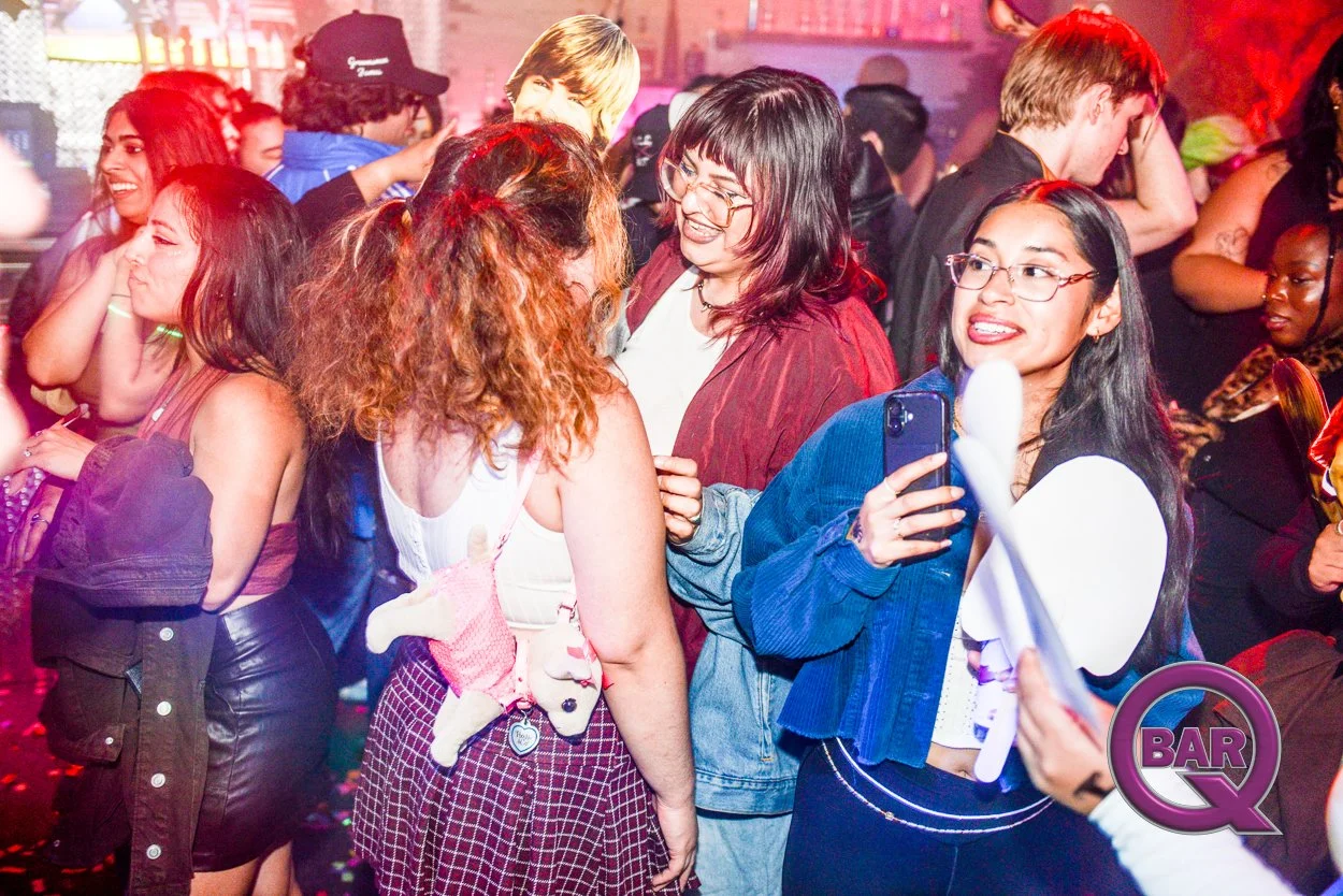 People dancing and socializing at a lively indoor bar or nightclub with colorful lighting, including women in casual and trendy outfits, some smiling and holding a phone, suggesting a fun night out.
