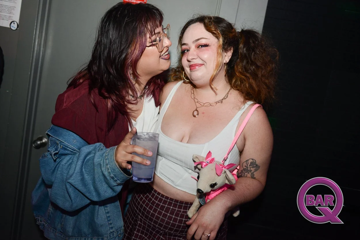 Two women at a social event, one holding a drink, smiling and wearing a white top with plaid pants, the other wearing glasses, a maroon jacket, and also smiling.