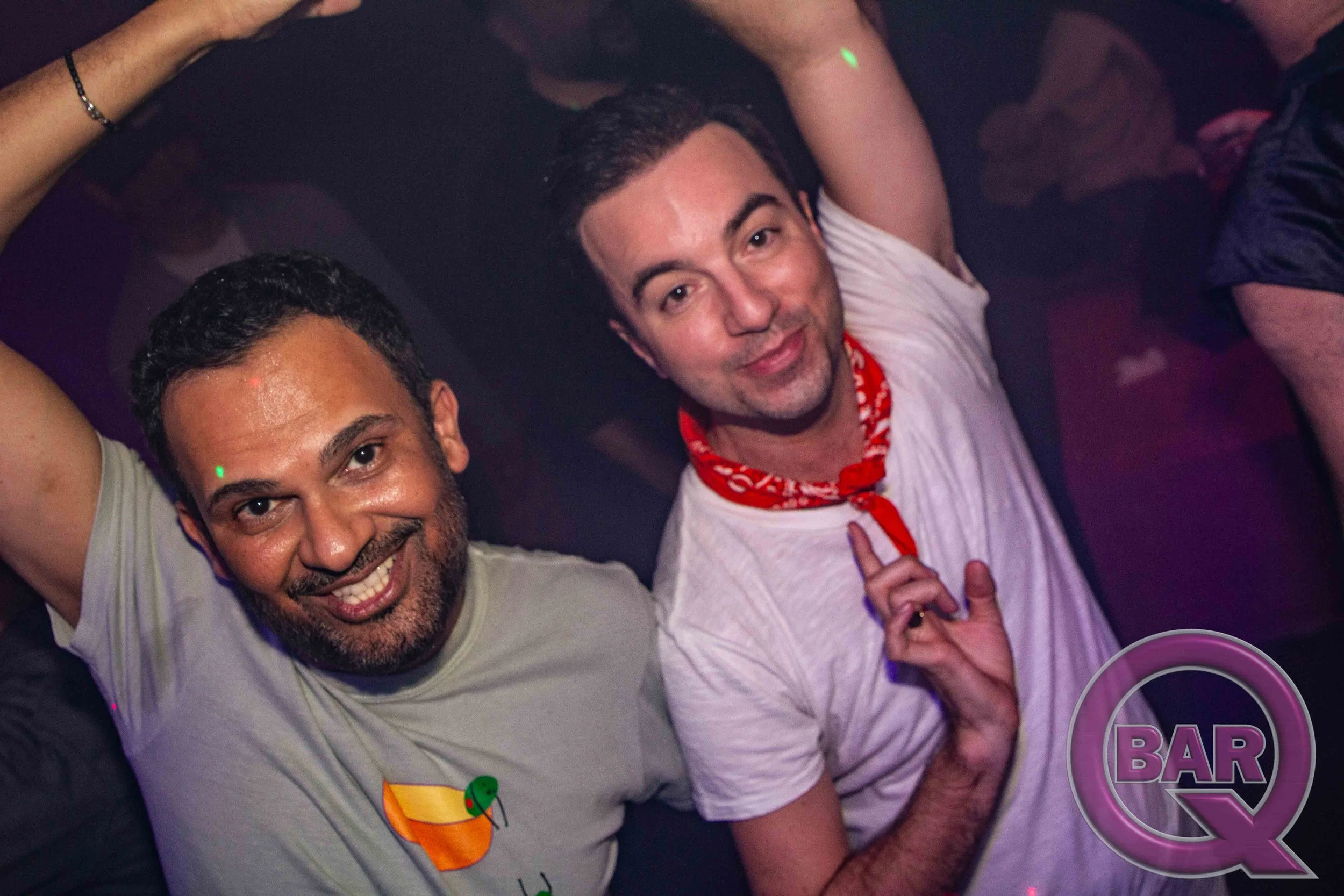 Two men at a party smiling and posing for a photo, with a woman partially visible behind them, in a dark setting with neon lights and drinks.