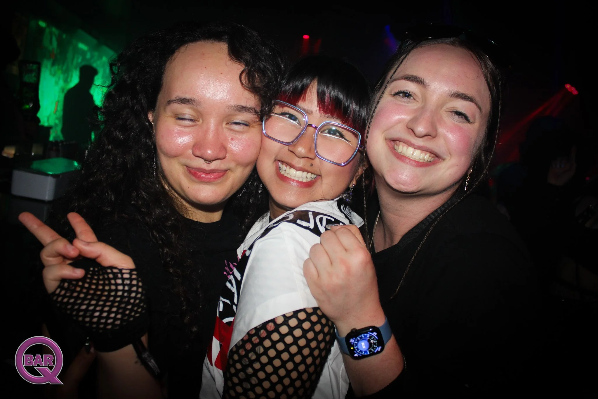 Three young women smiling and hugging in a dark club with colorful lights, with one wearing glasses and a smartwatch.