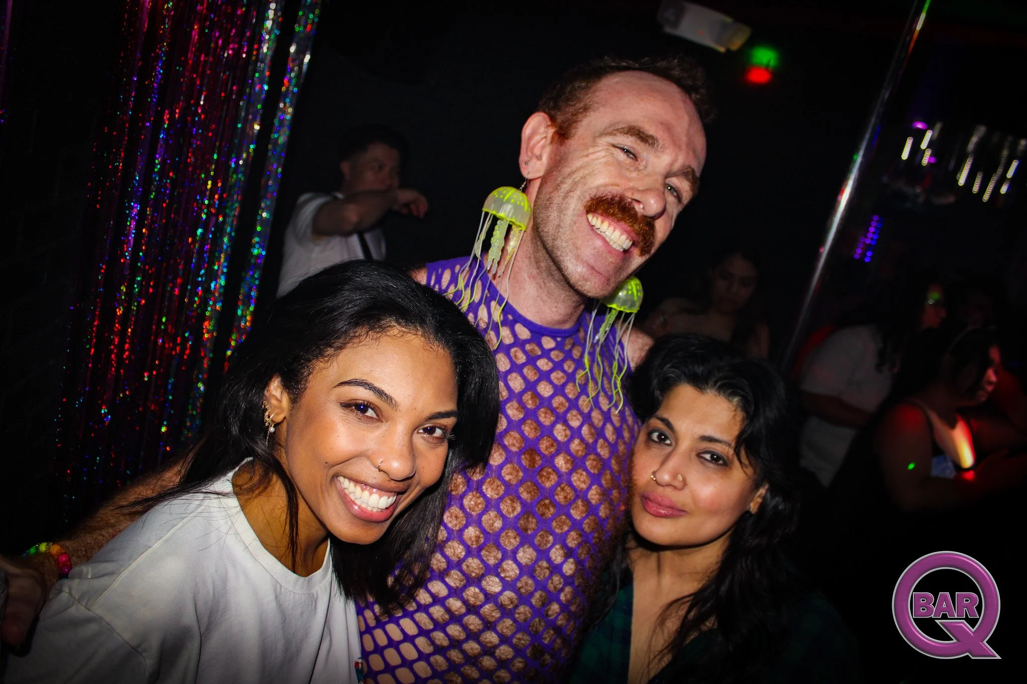 Group of four people smiling at a party, three women and one man with jellyfish earrings, in a dimly lit setting with colorful lights and a sticker with the word 'BAR' in the corner.