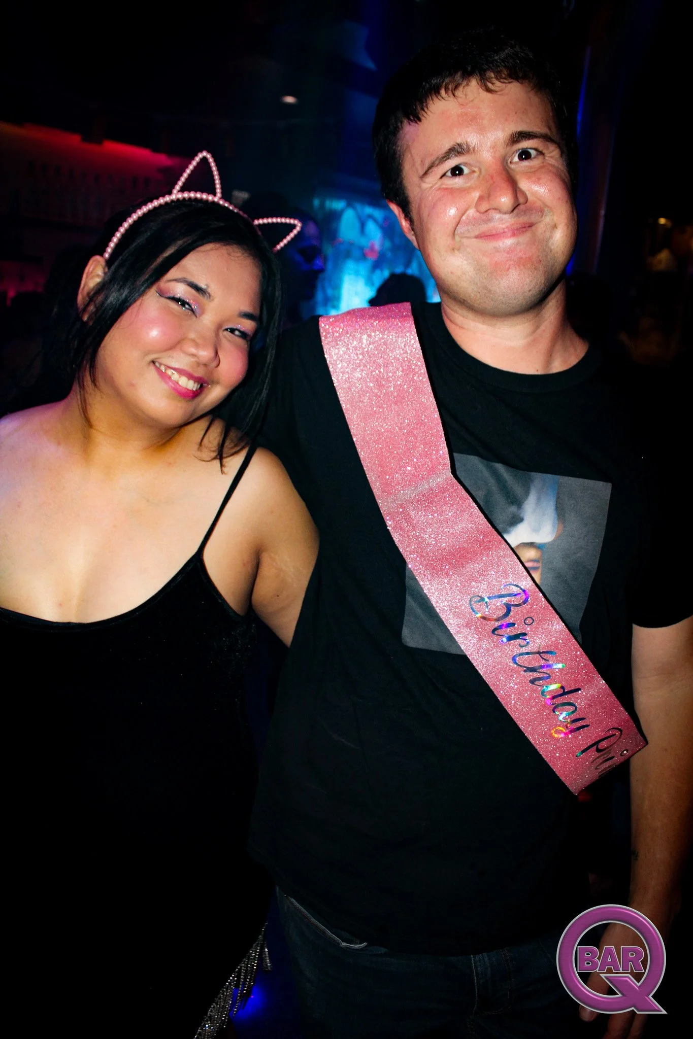 A woman wearing a black top and a pink headband shaped like cat ears is smiling, standing next to a man wearing a black t-shirt and a pink birthday sash, holding a round sign with a purple border that says 'BAR Q' in purple letters. They are in a dim