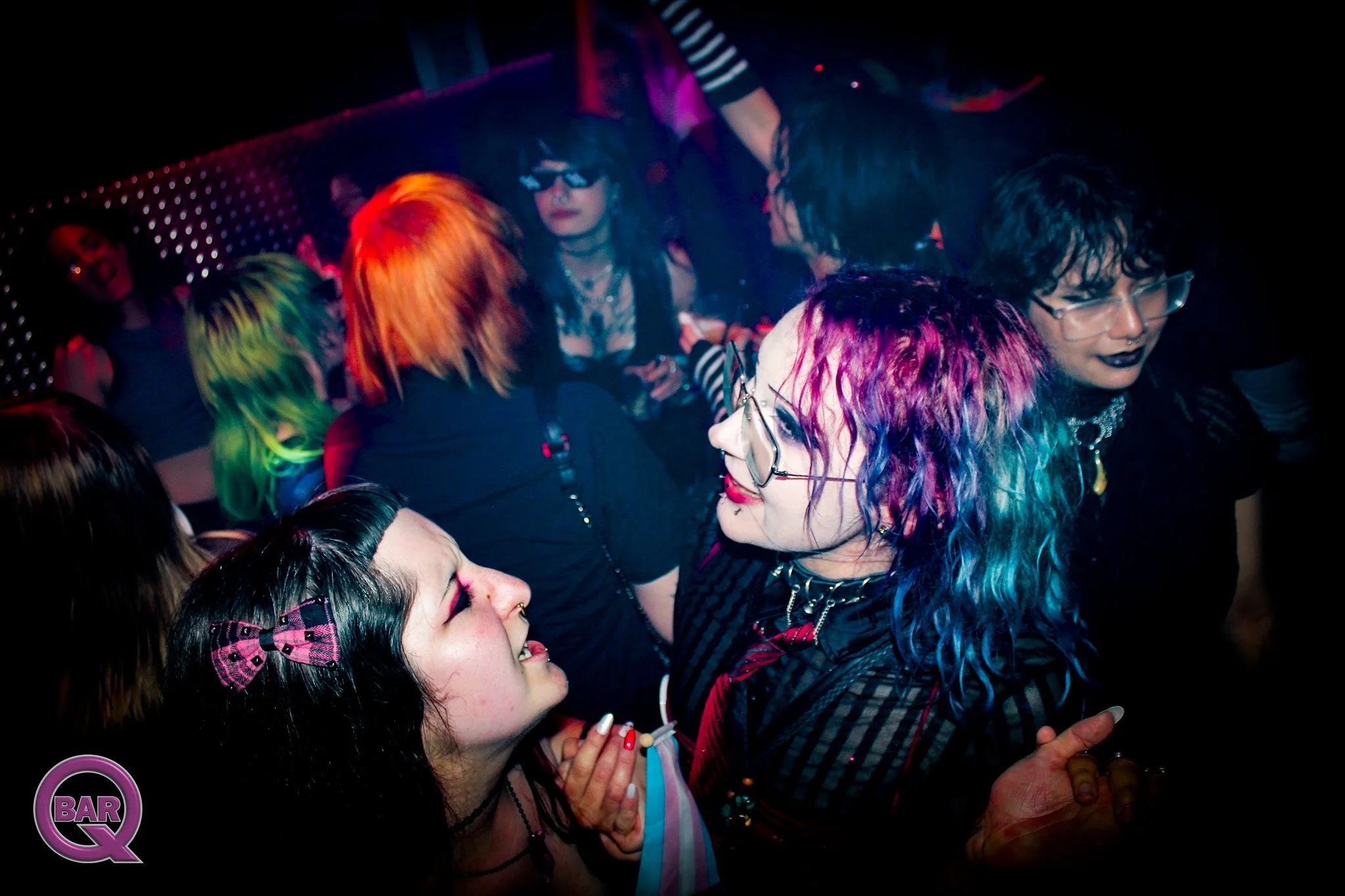 Group of young people with colorful hair and dark clothing dancing in a nightclub with neon lighting.