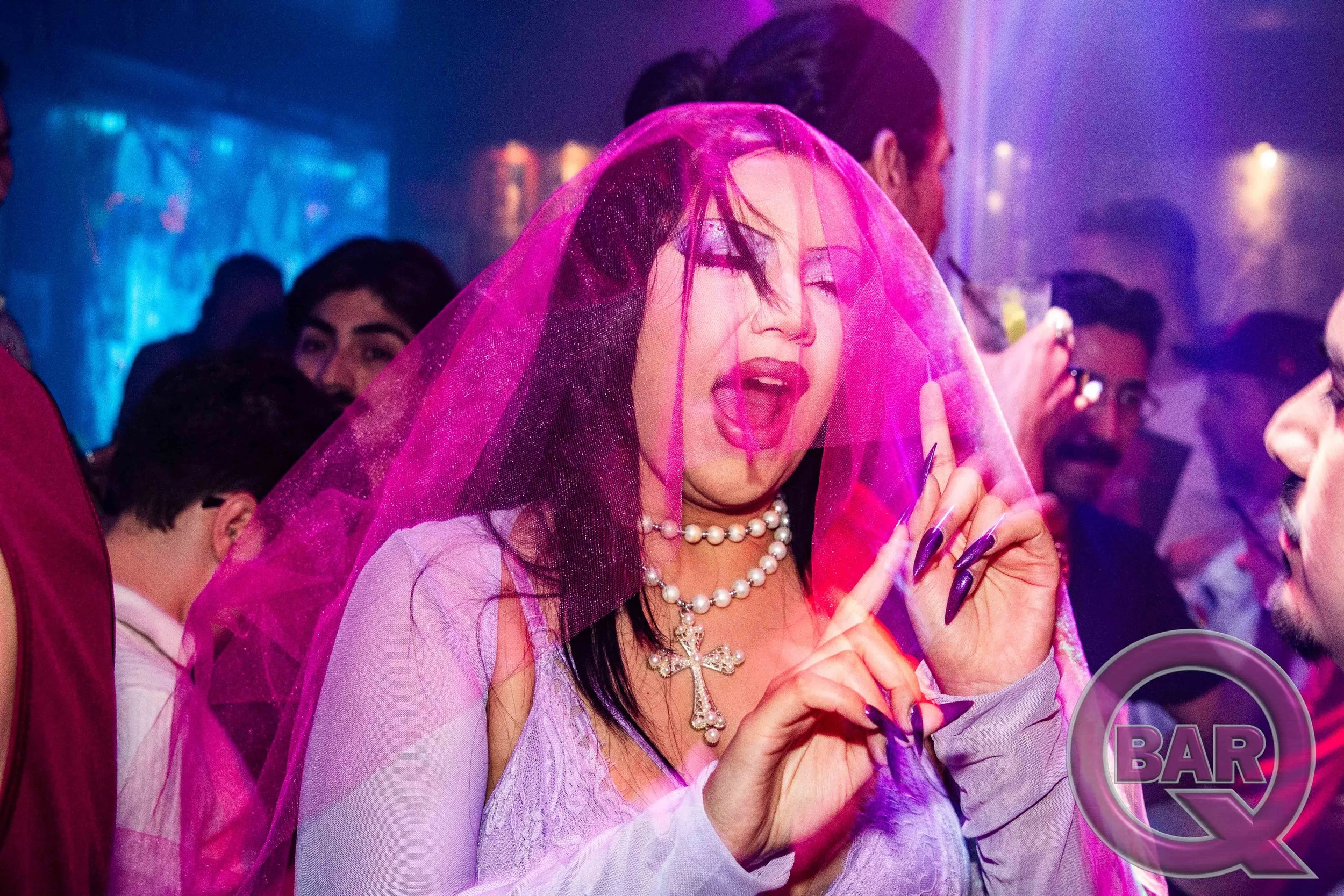 A woman with dark hair and pearl jewelry wearing a pink veil at a nightclub or party, surrounded by other people.