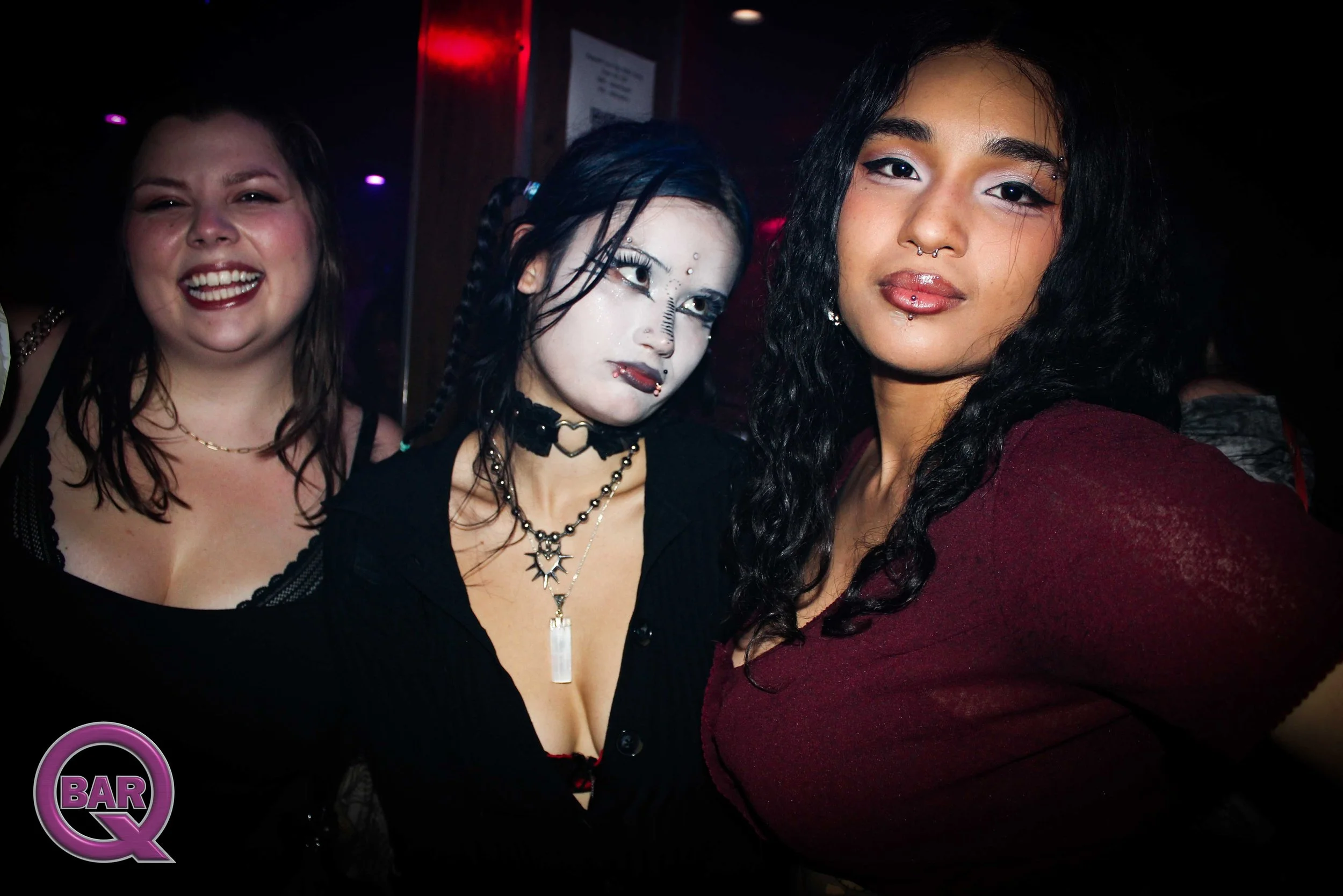 Three women posing for a photo at a bar, smiling and wearing dark clothing with gothic and alternative fashion styles. The woman in the middle has pale makeup, face piercings, and dark hair. The woman on the right has curly dark hair, a septum pierci