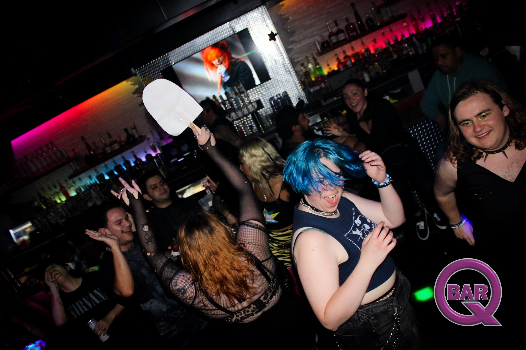 People dancing and enjoying themselves at a bar or club with colorful lighting, some with brightly dyed hair and alternative fashion styles. A bar shelf with bottles and a TV screen showing a performer are visible in the background.