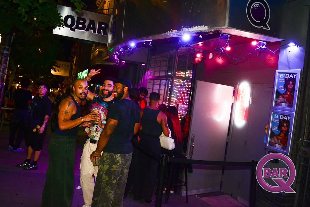 A group of friends smiling and taking a selfie outside a nightclub with neon signs and purple and pink string lights."QBAR" sign above the entrance and posters on the wall advertising 'B'DAY' celebration.