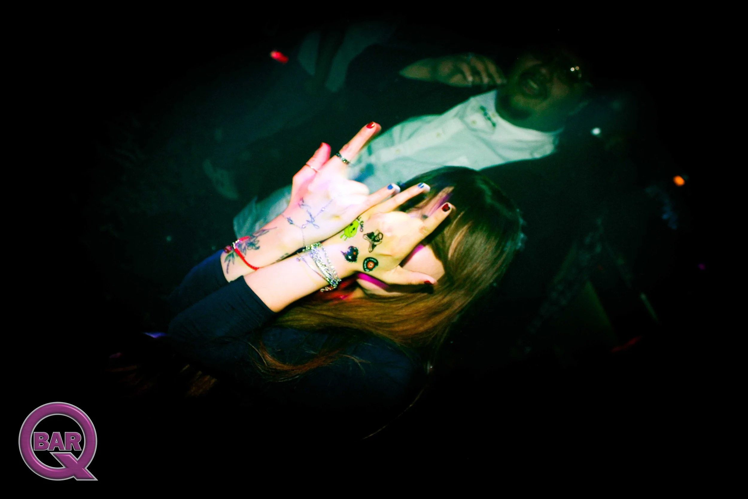 A woman with long hair and tattoos covering her hands and arms is in a dark setting, with a man wearing glasses and a light-colored shirt in the background. The woman is making a hand sign with her fingers.