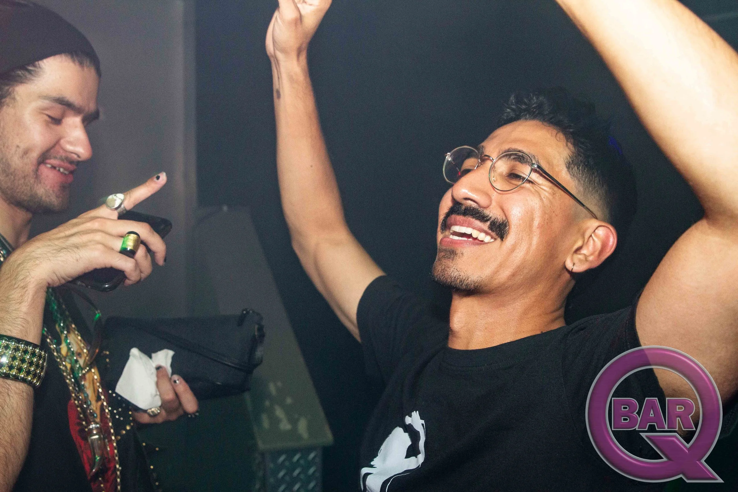 Two men smiling and looking at each other in a social setting. One is wearing glasses and a black shirt and the other is holding a phone and appears to be taking a picture or video.