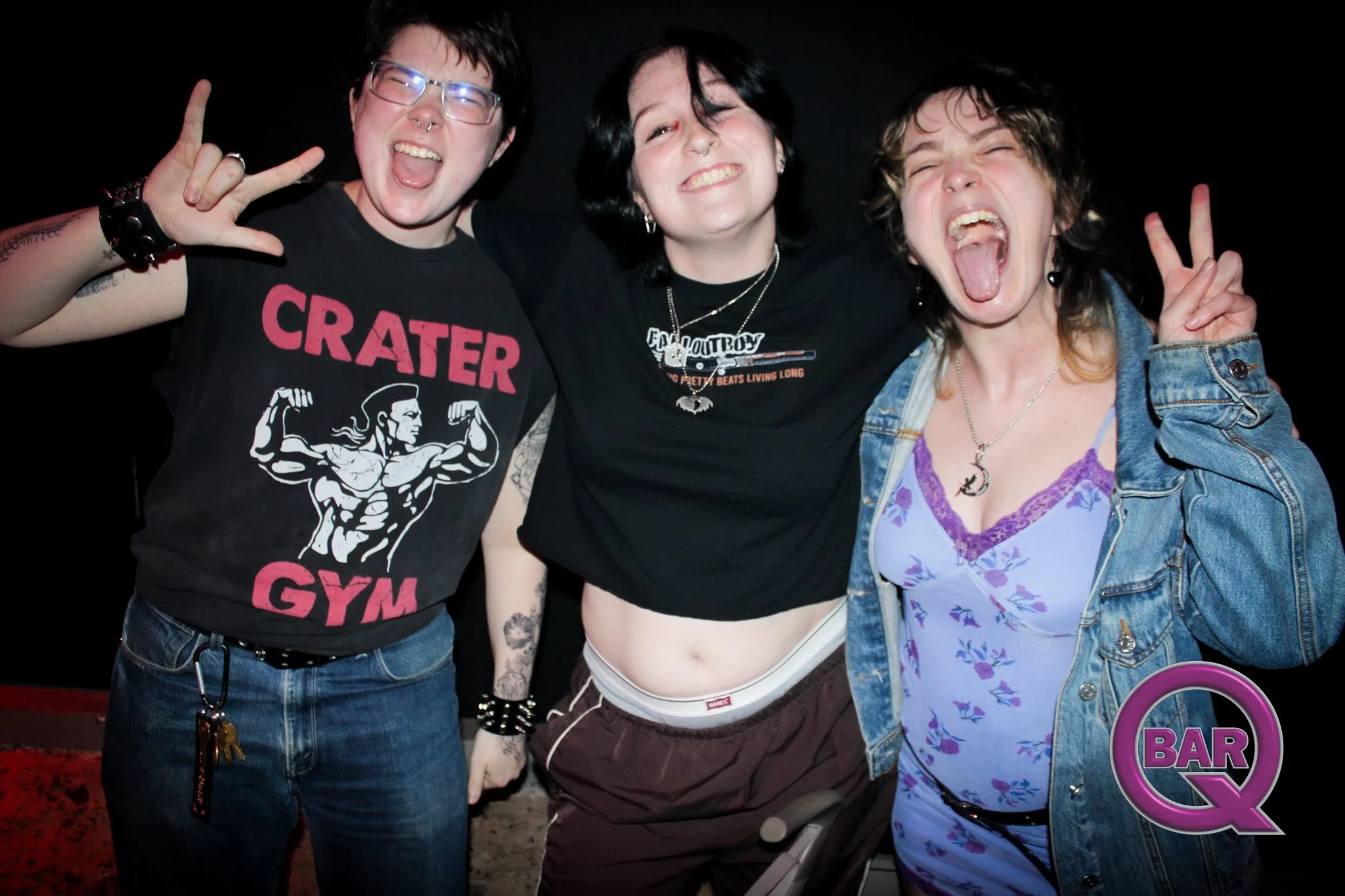 Three people posing at a bar, smiling and making rock and peace signs with their hands. The person on the left is wearing glasses, a black t-shirt with a graphic, and has bracelets. The person in the middle has dark hair, is wearing a black t-shirt w