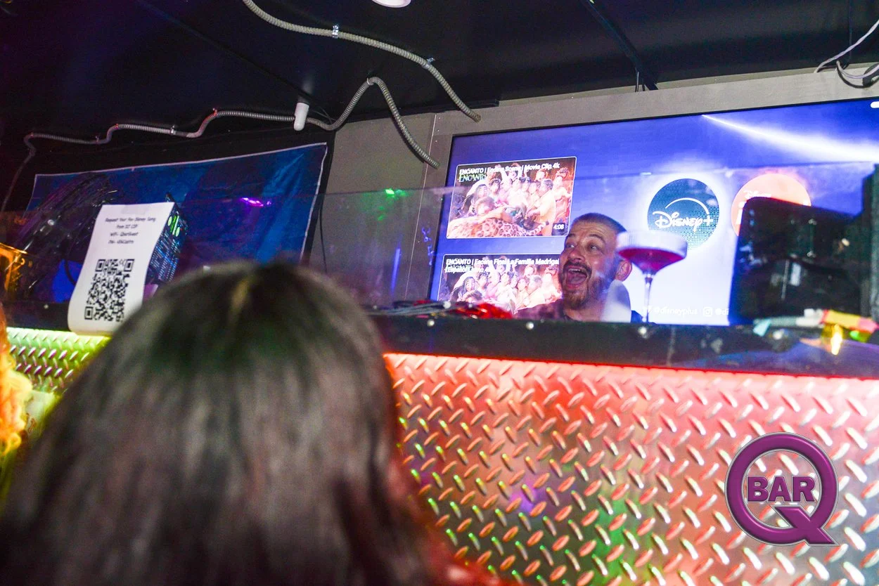 A man with a beard laughing behind a DJ booth at a bar, with a drink on the counter and a person with long hair in the foreground. There are two TV screens behind him displaying images, and the bar has a metallic textured counter with purple and gree