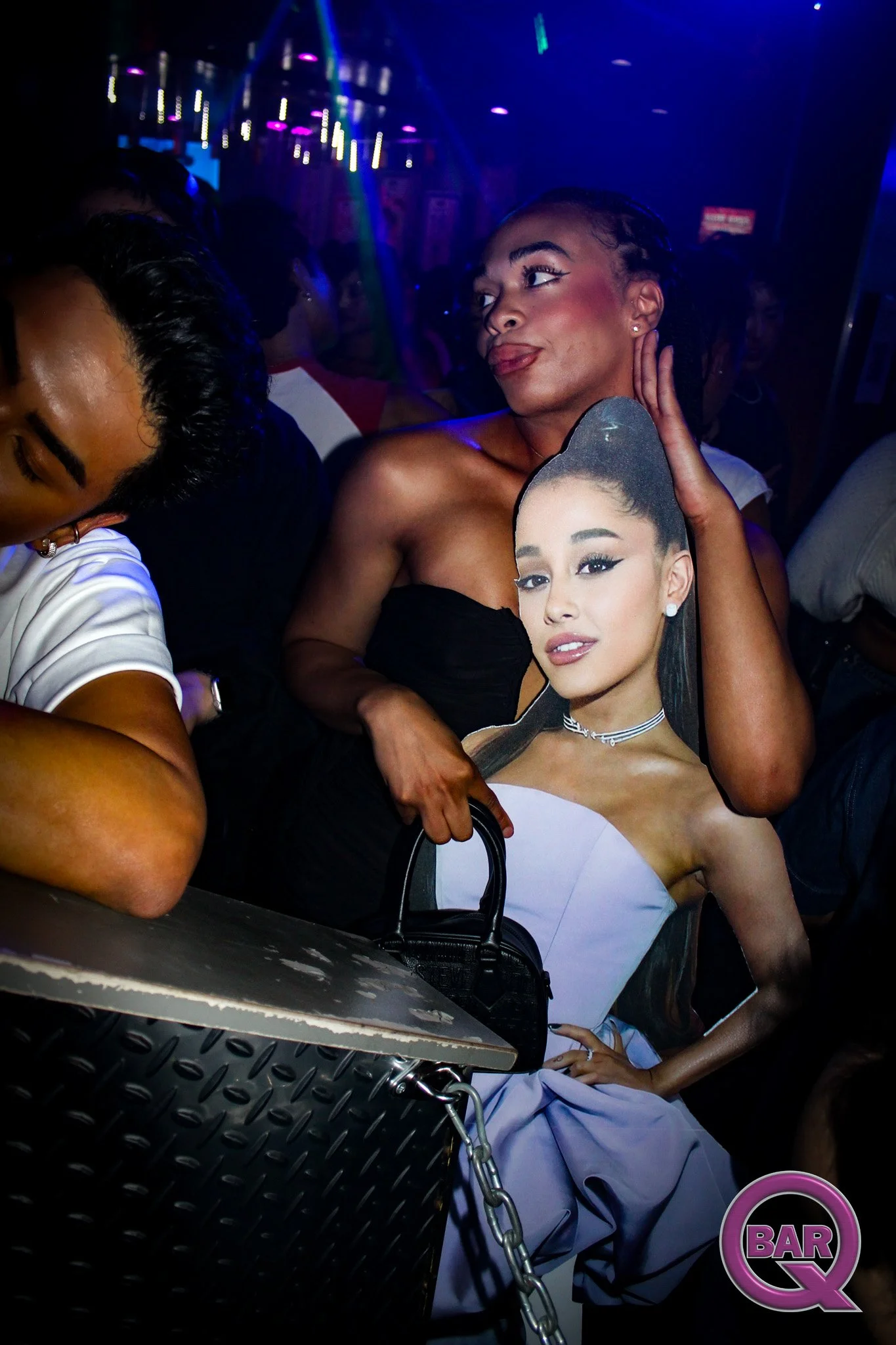 A woman with dark hair styled in a high ponytail is holding a cardboard cutout of another woman's face at a nightclub. The woman with the cutout is wearing a strapless black top, and the background features colorful lighting and other club patrons.