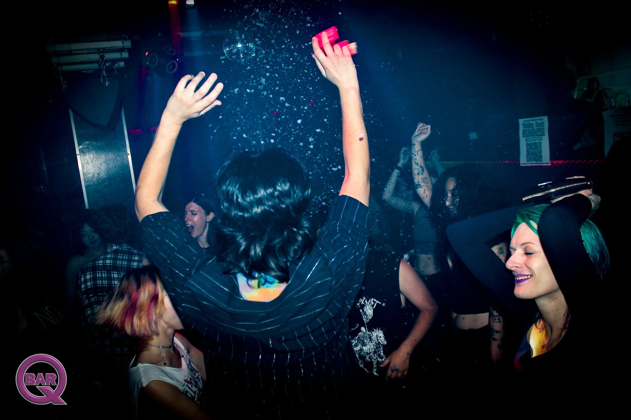 People dancing and celebrating in a nightclub, with one person raising their hands in the air. The scene is dark with colorful lighting and some confetti or water droplets in the air.