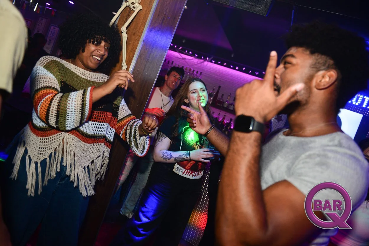 Four people are at a bar or nightclub, engaging in a lively conversation. A woman with curly hair and a colorful striped sweater is smiling and holding hands with a man with short, curly hair, who is gesturing with his thumb and wearing a smartwatch.