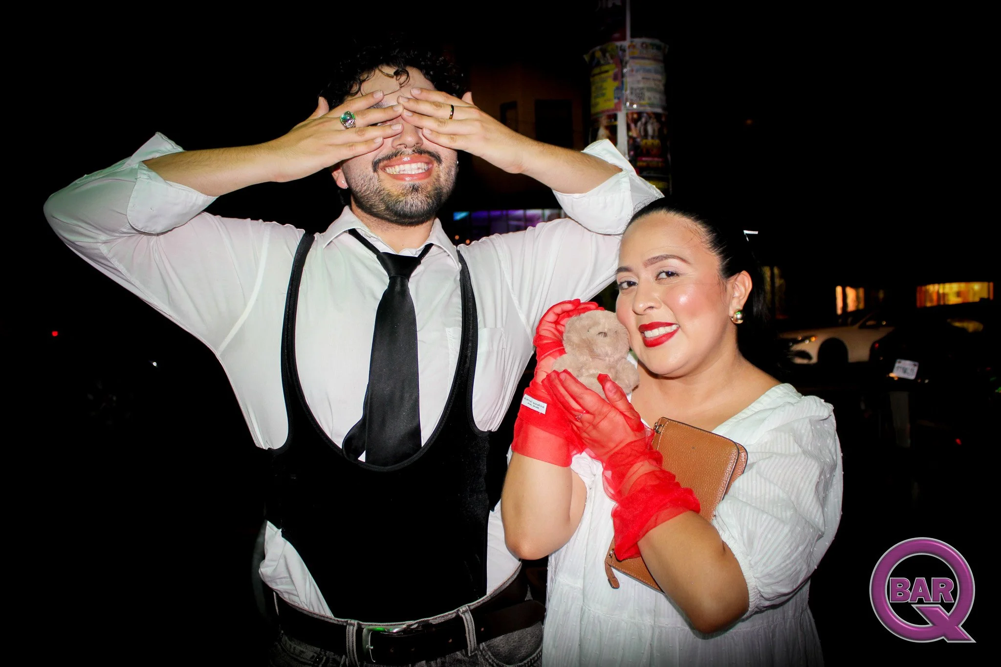 A man and woman smiling at the camera during a nighttime event. The man is dressed in a white shirt and black tie, with his hands covering his eyes, and has curly dark hair and facial hair. The woman is wearing a white outfit with red gloves, holding