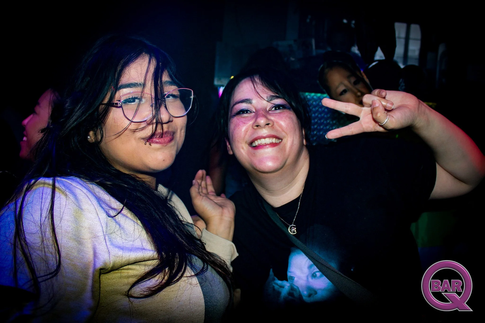 Two smiling women at a nightclub making peace signs, with other people in the background, dim lighting, and a neon sign.