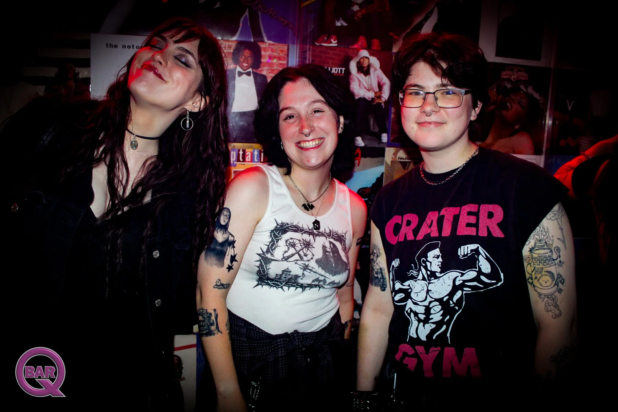 Three people smiling at a social event, with posters on the wall behind them. The person on the left has long dark hair, closed eyes, and shiny makeup. The person in the middle has short dark hair, tattoos on the arm, and is wearing a sleeveless shir