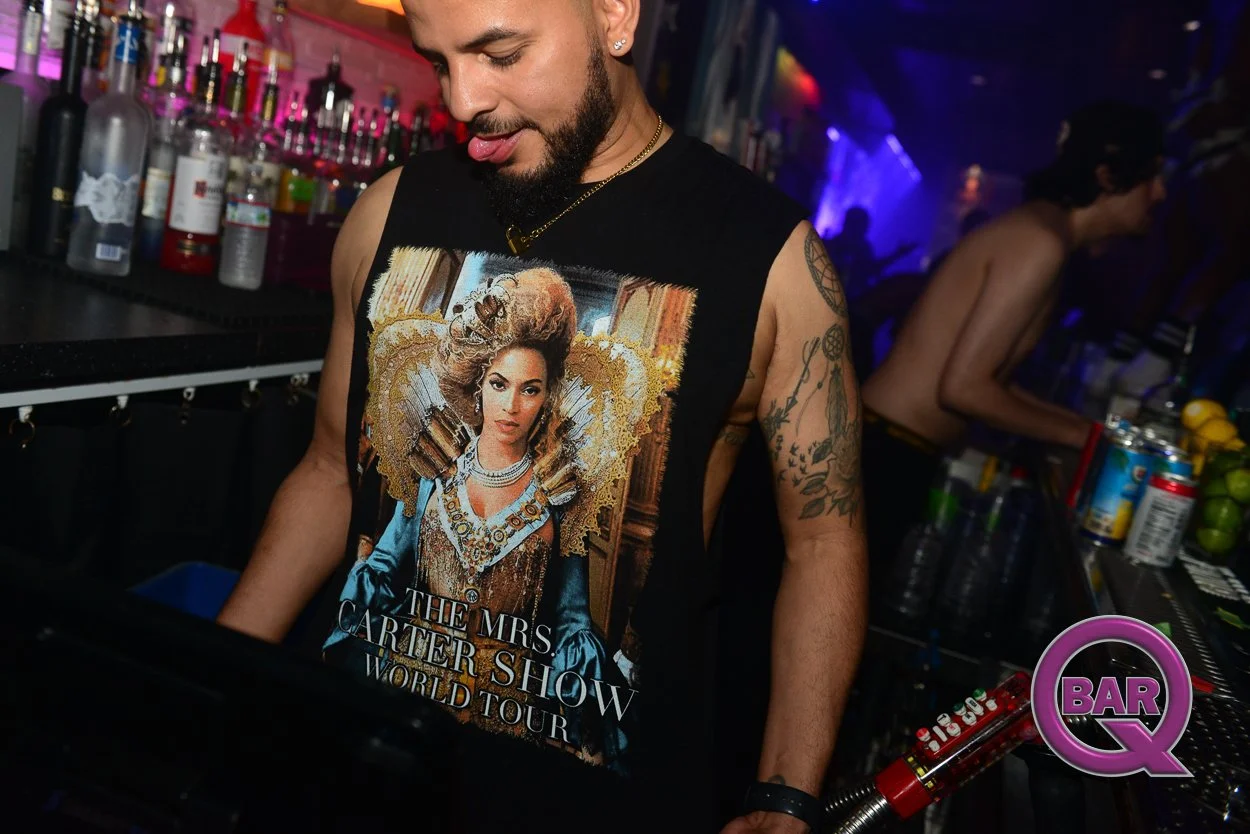 A male bartender with tattoos wearing a sleeveless graphic t-shirt, working behind a bar at a nightlife venue, with liquor bottles on shelves and colorful lighting in the background.