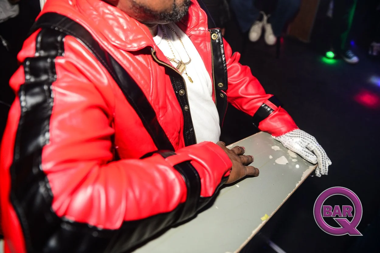 Person wearing a red and black jacket and white gloves playing at a bar or club, with a logo in the bottom right corner labeled 'BAR Q' and colorful lights in the background.