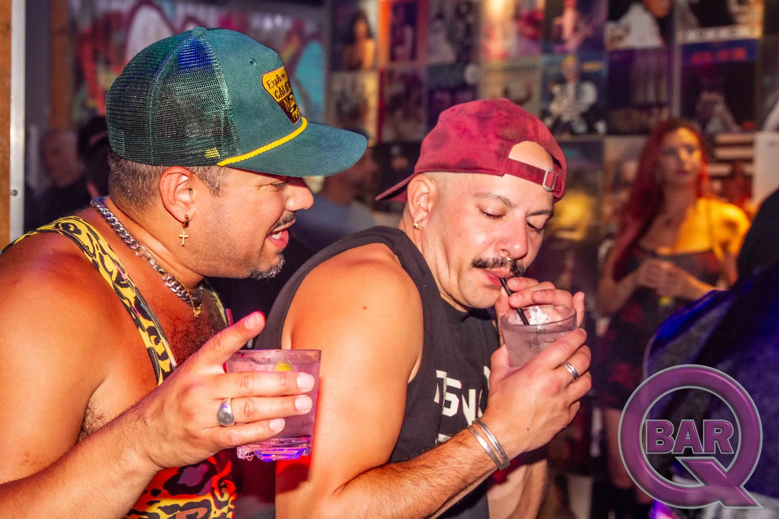 Two men sitting at a bar, each holding a drink, with a woman in the background. One man wears a green cap and jewelry, the other wears a red cap.
