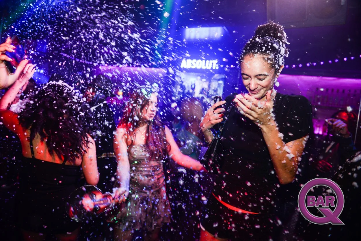 People dancing and celebrating in a nightclub with foam spraying in the air, colorful lights, and a sign that says 'ABSOLUT' in the background.