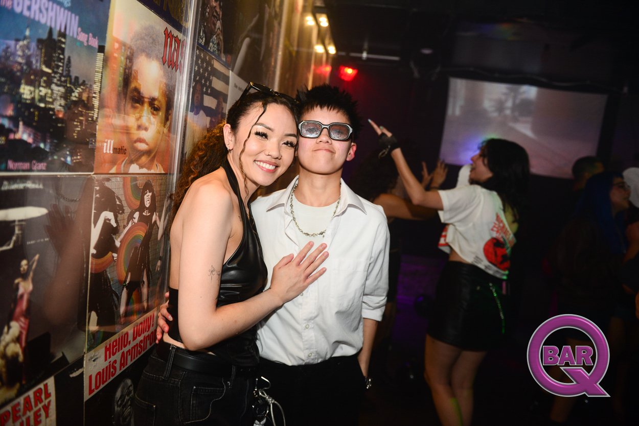 A young woman and a young man posing together at a nightclub, smiling. The woman is wearing a black sleeveless top, and the man is wearing sunglasses, a white shirt, and a chain necklace. The background shows other people dancing and a wall with post