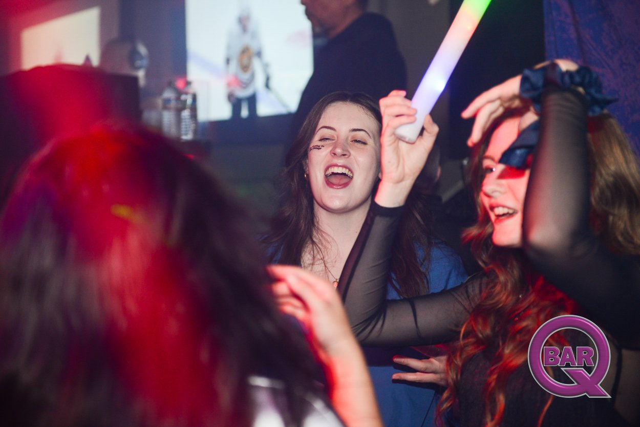 Group of women enjoying and dancing at a bar, with colorful lights and a screen in the background.