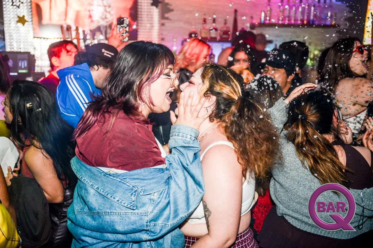 Two women with short dark hair and glasses wearing a denim jacket and a tank top, respectively, are happily sharing a kiss on a crowded dance floor with partygoers, confetti, and colorful lights in the background at a nightclub or bar.