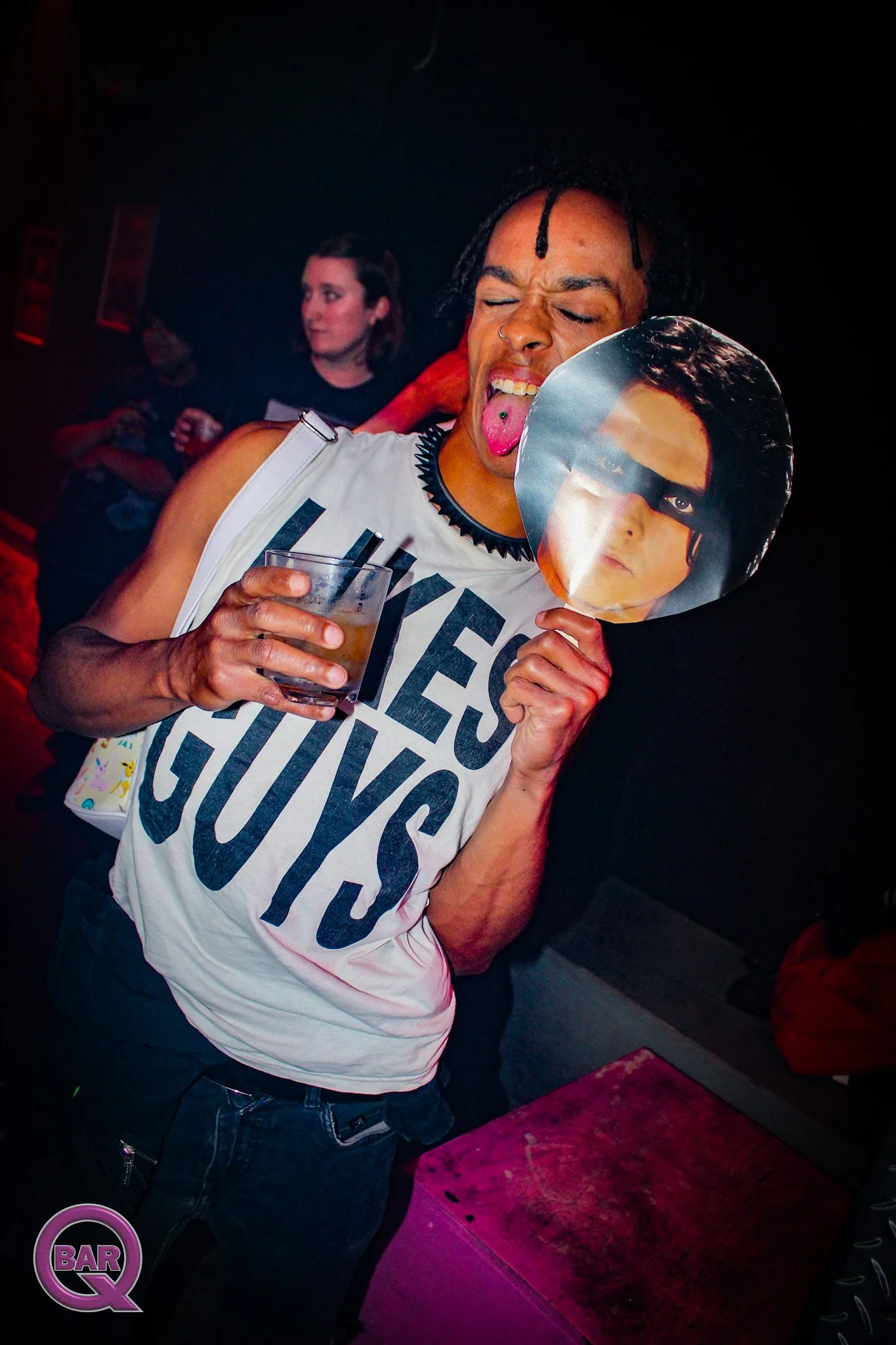 Person with dark skin, dreadlocks, and a nose ring holding a cutout of a person's face mask and a drink, at a dimly lit bar or club, with a black background and a purple BARQ logo in the bottom left corner.