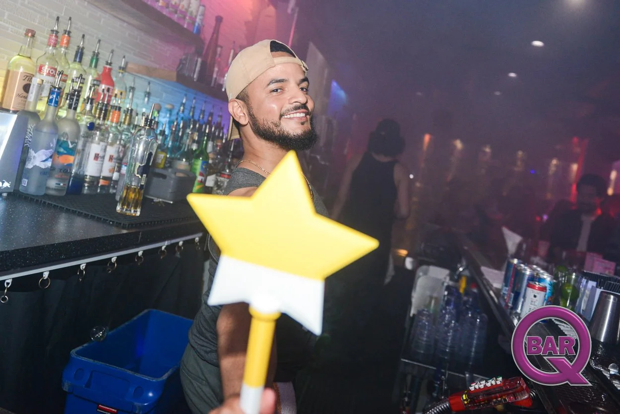 A smiling man with a beard and a backwards beige cap behind a bar counter, holding a yellow star-shaped wand, in a dimly lit bar or nightclub with colorful bottles and a woman in the background.