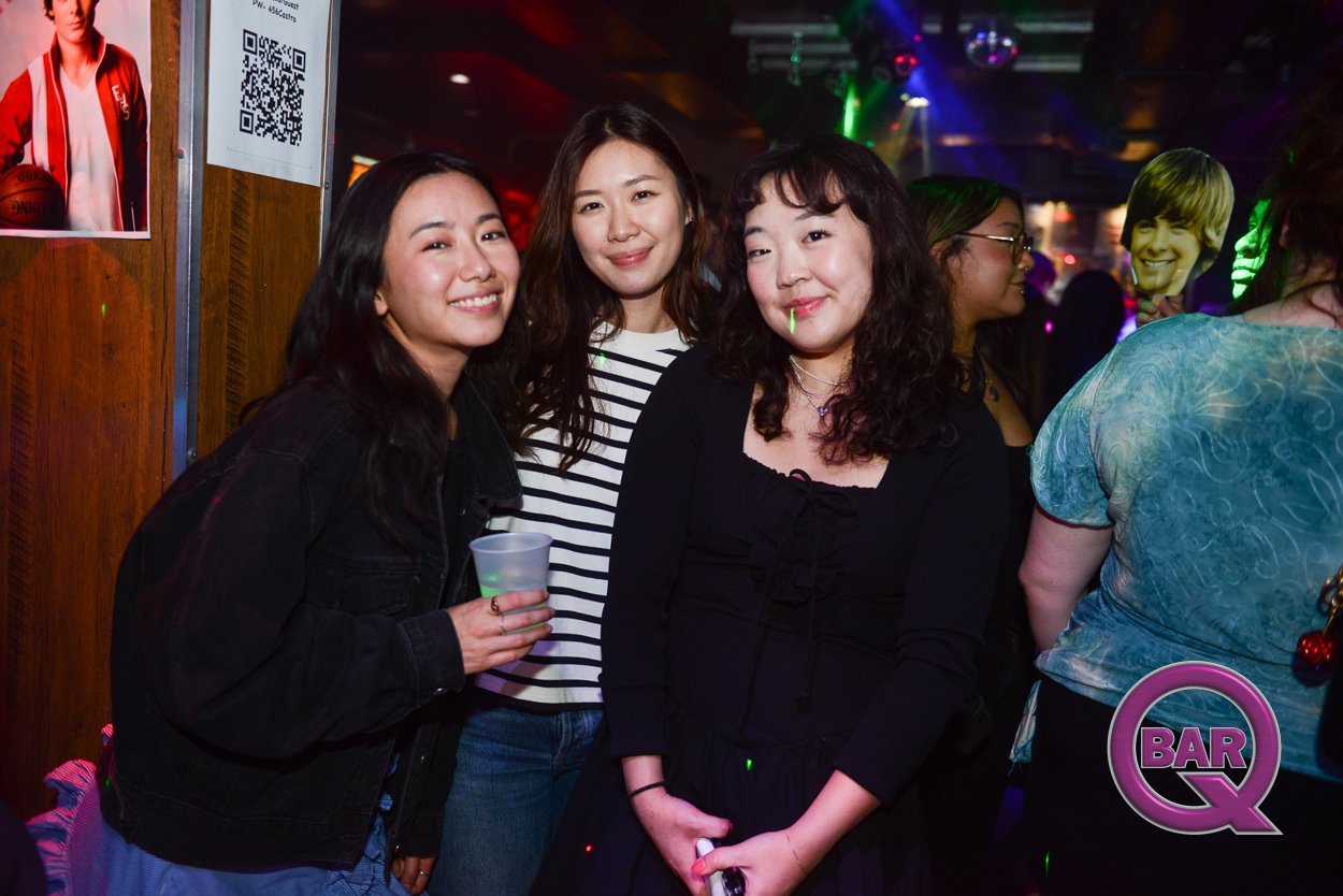 Three women smiling and enjoying themselves at a bar, with colorful lights and other people in the background.