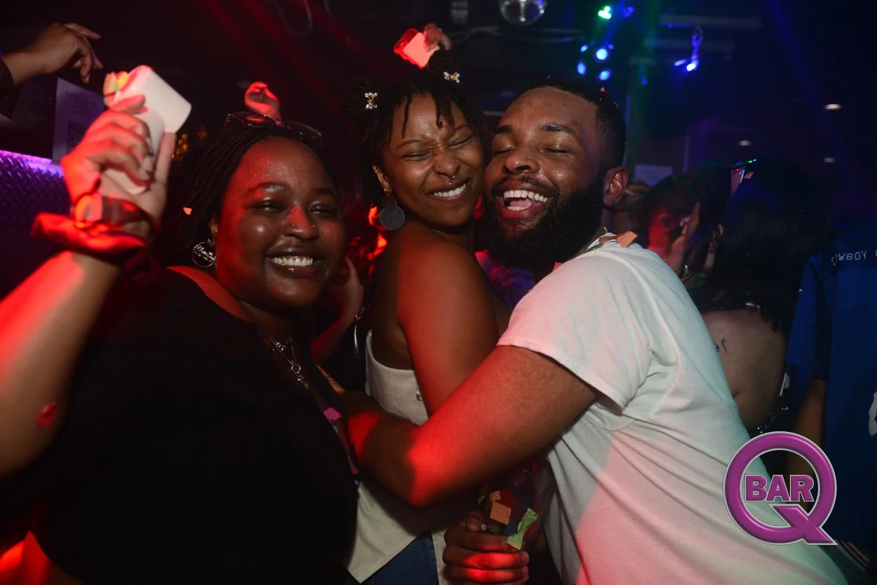 Three smiling people dancing and having fun at a nightclub with colorful lights and a 'BarQ' logo in the bottom right corner.