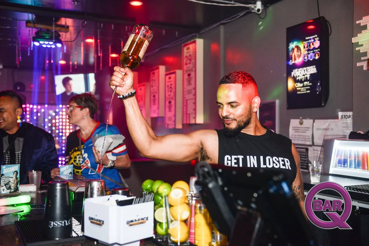 A man with a beard and short hair at a bar, wearing a black sleeveless shirt that reads 'GET IN LOSER', holding an upside-down bottle of beer. Two people are in the background, a woman with glasses and a man, both smiling. The bar counter has lemons,