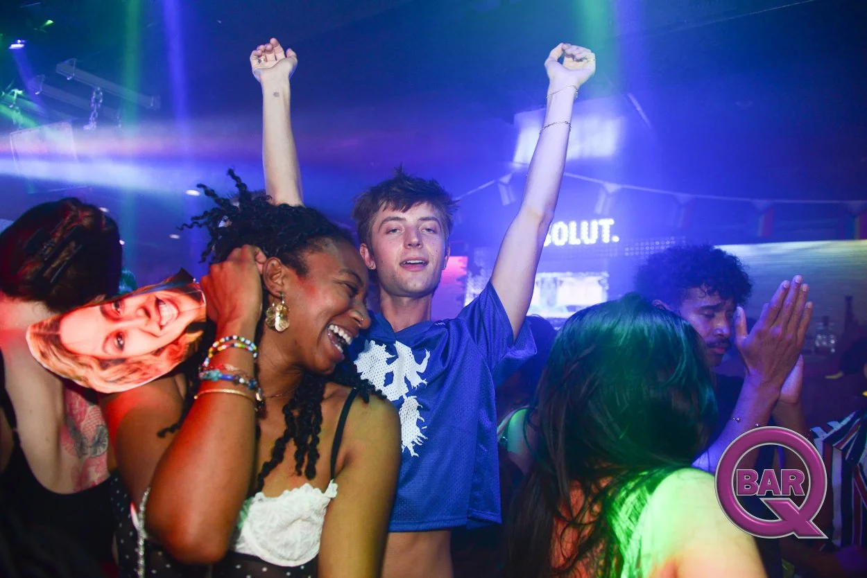 People dancing and celebrating at a nightclub with colorful lights, smiles, and raised hands.