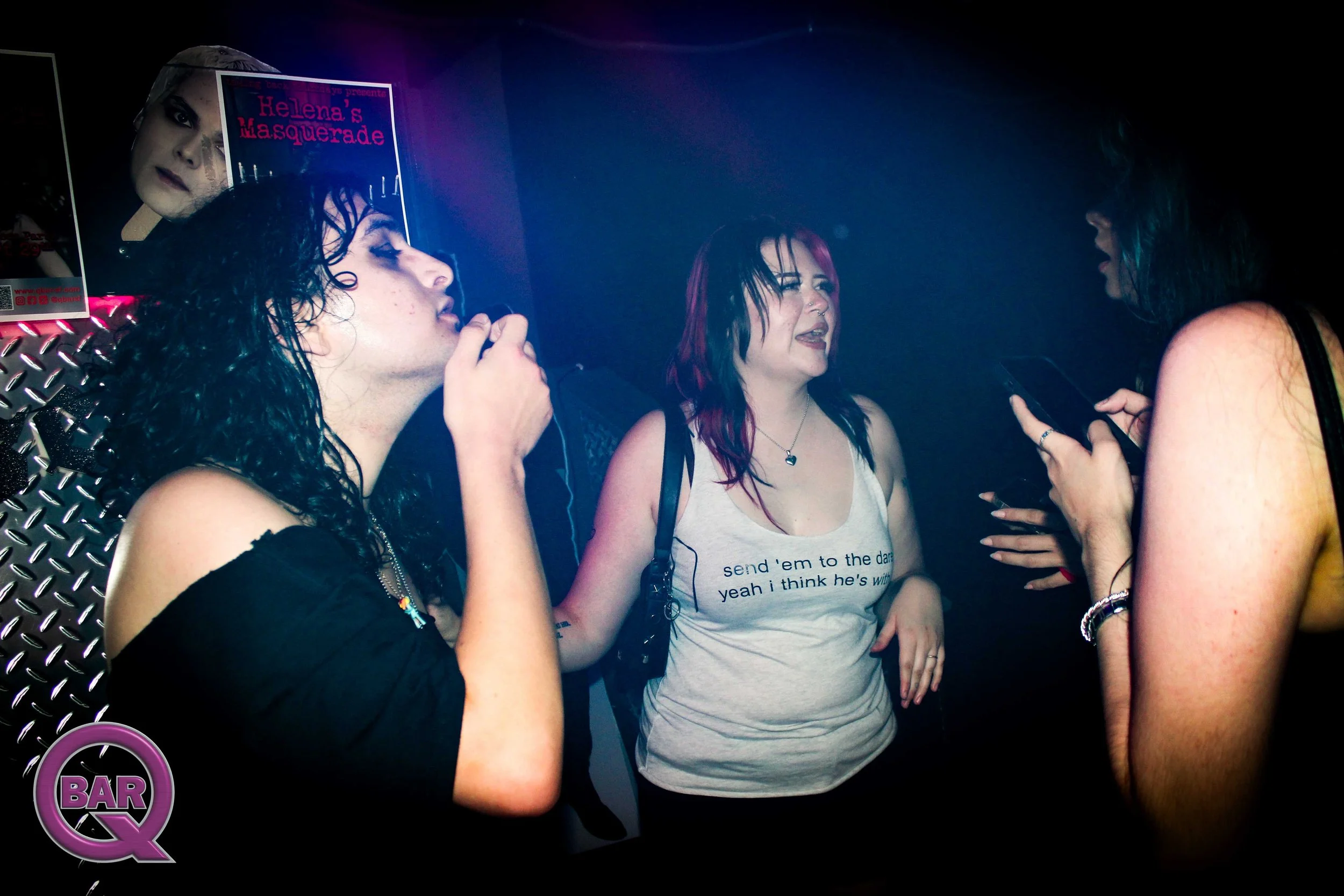 Group of women with dark hair, one with pink tips, talking at a bar, with posters on the wall behind them that include one advertising Helena's Masquerade event, and a neon sign that says BQ.