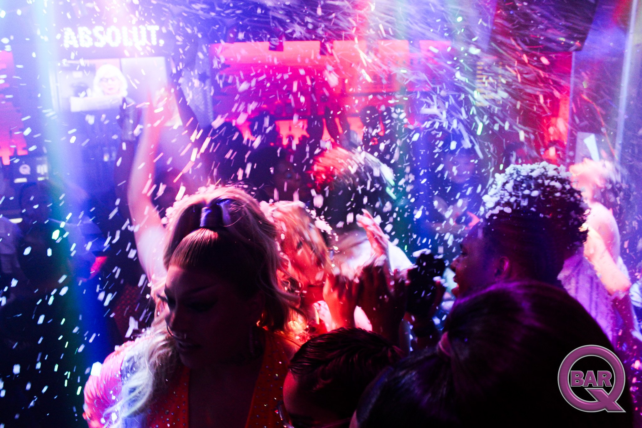 People celebrating at a party with confetti falling, colorful lights, and a bar sign visible in the background.