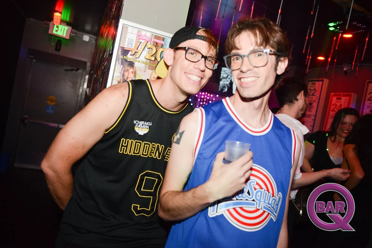 Two men with glasses posing together at a bar or nightclub, one wearing a black basketball jersey and the other wearing a blue basketball jersey, with colorful neon lights and other people in the background.