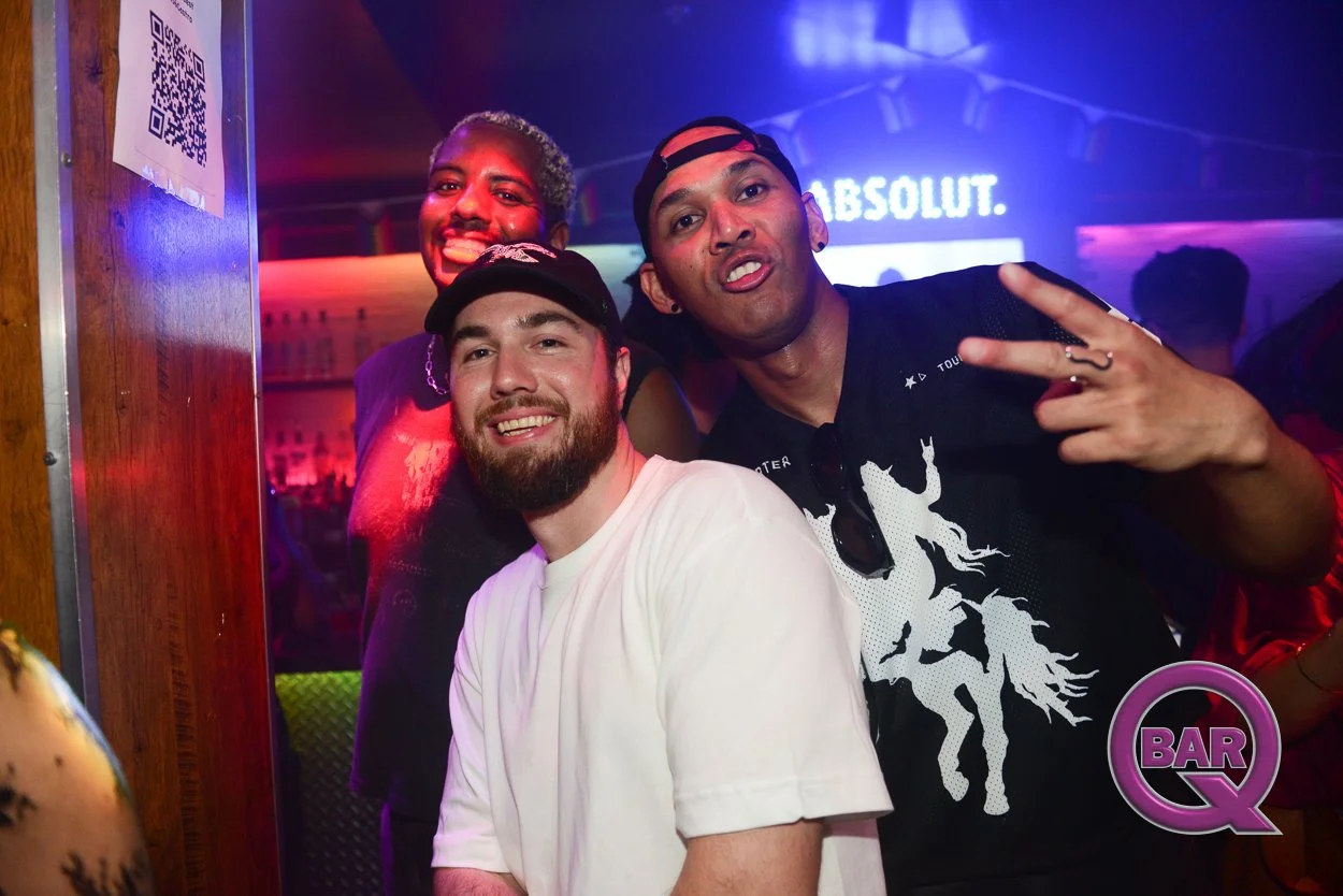 Three smiling men posing together in a nightclub with colorful lights, one in a white t-shirt and cap, one making a peace sign, and the third smiling behind them.